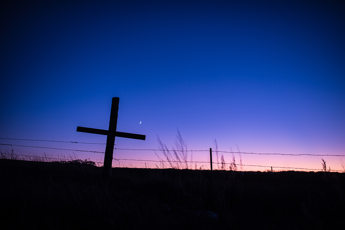 beautiful shot of a cross at sunset with the moon and fence in the background